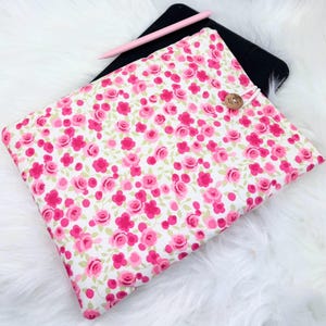 May include: A floral tablet sleeve with a pink rose pattern on a white background. The sleeve has a wooden button closure and a pink stylus resting on top. The sleeve is designed to fit a tablet.