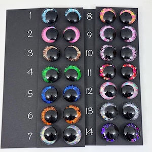 May include: A collection of fourteen pairs of round, glossy, black cabochons with colorful glitter accents. The glitter colors include blue, pink, brown, green, orange, and purple. Each pair is displayed on a black card with a number from 1 to 14.
