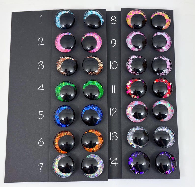 May include: A collection of fourteen pairs of round, glossy, black cabochons with colorful glitter accents. The glitter colors include blue, pink, brown, green, orange, and purple. Each pair is displayed on a black card with a number from 1 to 14.