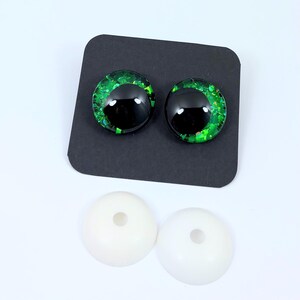 May include: Two round, black and green glitter craft eyes with white backing pieces. The eyes have a black pupil and a green glitter iris. The backing pieces are white and have a small hole in the center.