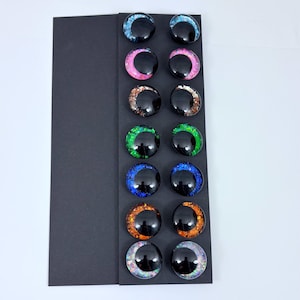 May include: A collection of twelve glossy, black, round glass eyes with colorful glitter accents. The glitter colors include blue, pink, copper, green, blue, orange, and iridescent. The eyes are arranged in rows on a black card.