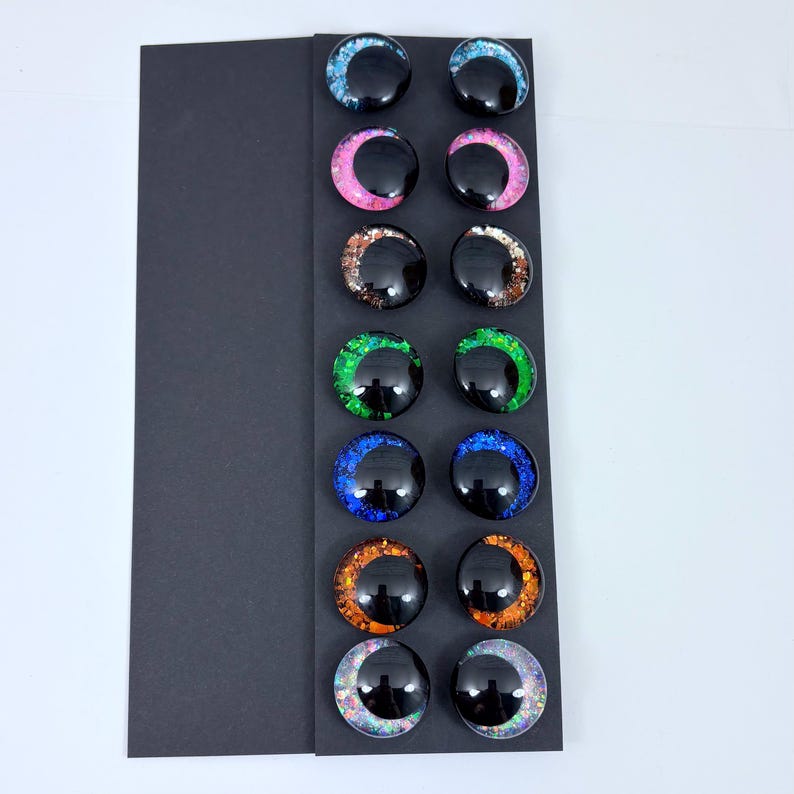May include: A collection of twelve glossy, black, round glass eyes with colorful glitter accents. The glitter colors include blue, pink, copper, green, blue, orange, and iridescent. The eyes are arranged in rows on a black card.