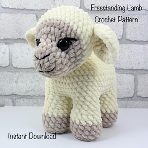 May include: A crocheted lamb in cream and grey, with large black eyes and a smiling expression. The text reads "Freestanding Lamb Crochet Pattern" and "Instant Download". The lamb is a soft toy.