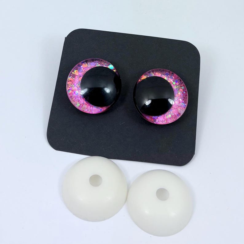 May include: Two black and pink crescent-shaped doll eyes with glitter accents sit on a black square. Below are two white, round doll eye backs with a hole in the center. Craft supplies.