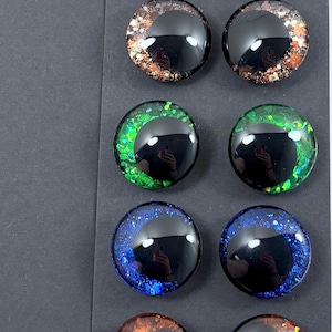 May include: A collection of twelve glossy, round, glass eye cabochons in various colors. The eyes feature a black pupil and colorful, glittery irises in shades of blue, pink, green, brown, orange, and silver. The cabochons are arranged in rows of two.