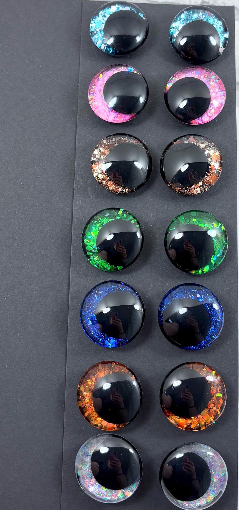 May include: A collection of twelve glossy, round, glass eye cabochons in various colors. The eyes feature a black pupil and colorful, glittery irises in shades of blue, pink, green, brown, orange, and silver. The cabochons are arranged in rows of two.