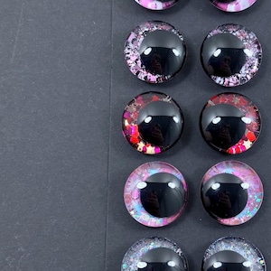 May include: A collection of twelve glossy, round, black-centered craft eyes with colorful, glittery surrounds. The eyes feature various color schemes, including pink, red, purple, and blue, with glitter accents. They are arranged in pairs on a dark gray surface.