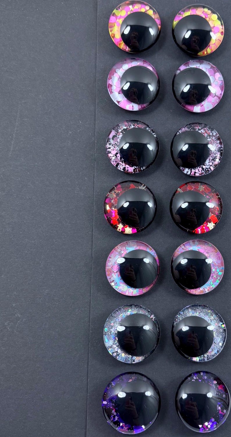 May include: A collection of twelve glossy, round, black-centered craft eyes with colorful, glittery surrounds. The eyes feature various color schemes, including pink, red, purple, and blue, with glitter accents. They are arranged in pairs on a dark gray surface.