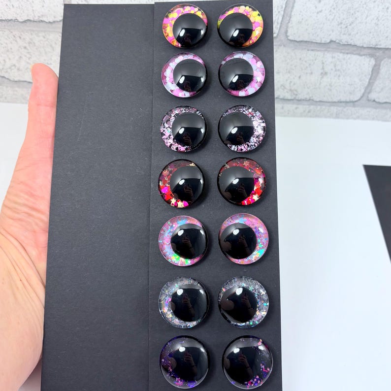 May include: A collection of twelve glossy, black, round craft eyes with colorful, glittery irises. The irises feature pink, purple, yellow, and red hues. The eyes are arranged on a black card.