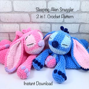 May include: Two crocheted alien plush toys in pink and blue, resembling a bunny, with closed eyes. The pink plush has lavender accents, while the blue plush has pink accents. The text "Sleeping Alien Snuggler 2 in 1 Crochet Pattern" and "Instant Download" are visible.