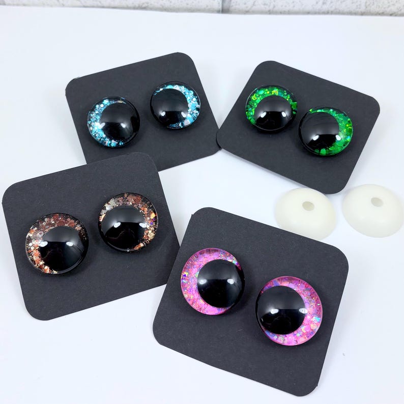 May include: A collection of doll eyes in various colors, including blue, green, brown, and pink, each with a black pupil. The eyes are displayed on small black cards, with two white backing pieces visible.
