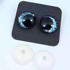 May include: Two glass eye cabochons with a black iris and blue and white accents, displayed on a black surface. Below are two white, round, plastic eye bases with a hole in the center. These craft supplies are for doll making or other art projects.