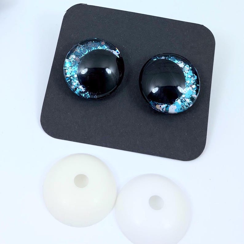 May include: Two glass eye cabochons with a black iris and blue and white accents, displayed on a black surface. Below are two white, round, plastic eye bases with a hole in the center. These craft supplies are for doll making or other art projects.