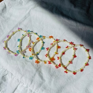 May include: A collection of beaded bracelets with colorful floral and fruit designs. The bracelets feature white beads with accents of purple, blue, orange, and red. The bracelets are arranged on a white fabric surface.
