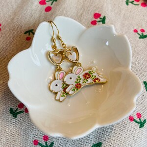 May include: Gold dangle earrings featuring a white rabbit with floral details and a heart-shaped charm. The rabbit is depicted with pink ears and red flowers.