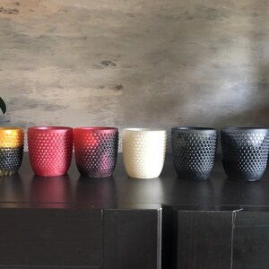 May include: Six textured candle holders in different colors, including gold, red, black, and white. The holders are arranged in a row on a black surface.