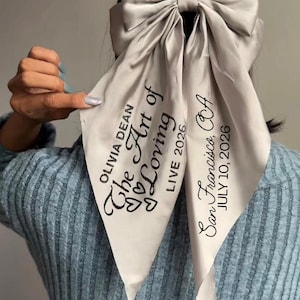 May include: A silver satin hair bow with long, flowing tails. The tails are printed with the text "OLIVIA DEAN The Art of Loving LIVE 2026" and "San Francisco, CA JULY 10, 2026". The bow is tied at the back of the head.