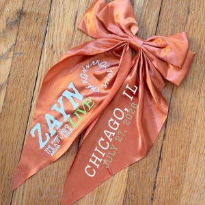 Op de afbeelding: Een oranje satijnen haarstrik met lange staarten. De strik heeft de tekst "ZAYN LIVE" in wit en groen, en "CHICAGO, IL JULY 27, 2026" in wit. De strik ligt op een houten oppervlak.