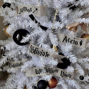 May include: A white artificial Christmas tree decorated with black and gold ornaments and burlap ribbon banners with Harry Potter spells written in black. The spells include "Lumos", "Accio", "Obliviate", "Riddikulus", "Wingardium Leviosa".
