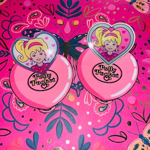 Może przedstawiać: Dwa różowe kompakty Polly Pocket z wieczkami w kształcie serca, każdy z blond dziewczynką z kucykiem. Kompakty znajdują się na żywym różowym tle z motywami motyli i kwiatów. Na każdym kompakcie wydrukowano napis "Polly Pocket".