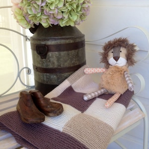Puede incluir: Una escena decorativa con un juguete de león de peluche, una manta de punto con un diseño de tablero de ajedrez y un recipiente de metal vintage que contiene un ramo de flores verdes y rosas. También hay dos hormas de zapatos de madera.