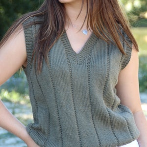 May include: Olive green knitted vest with a V-neck. The sleeveless top has vertical ribbed detailing and a slightly flared hem. The vest is made of a soft, textured knit fabric. The model is wearing white trousers.