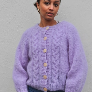 May include: A light purple, cable-knit cardigan with wooden buttons. The sweater has a round neckline and long sleeves. The model is wearing blue jeans. The cardigan is a soft, fuzzy knit.