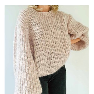 Może przedstawiać: Różowy sweterek z dzianiny z okrągłym dekoltem i długimi rękawami. Sweter ma luźny krój i jest wykonany z miękkiej, puszystej przędzy. Sweter jest pokazany na modelce w czarnych spodniach. Na dole zdjęcia widnieje tekst "#159 ISLA JERSEY".