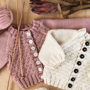 May include: A close-up shot of two hand-knitted baby cardigans in soft pink and cream colors. The pink cardigan has white buttons and a textured pattern, while the cream one has dark brown buttons. Knitting needles and yarn balls are also visible.