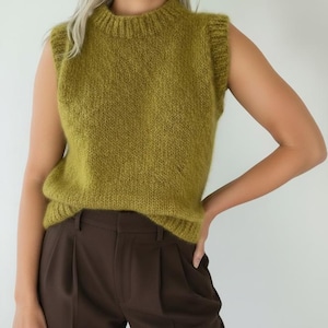 May include: A sleeveless knit sweater vest in a vibrant olive green color featuring a ribbed mock neck collar and ribbed armholes. The garment has a soft, fuzzy texture and is paired with dark brown trousers that have visible front pleats.