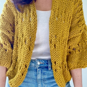May include: A mustard yellow knitted cardigan with a loose fit and open front. The cardigan has a delicate stitch pattern and is shown worn over a white vest top and blue jeans.