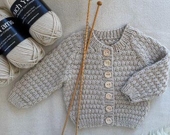 May include: A light beige, hand-knitted baby cardigan with wooden buttons and knitting needles. Two skeins of Touch Yarns merino wool are visible. The cardigan is approximately 30 cm long.