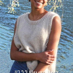 Pode incluir: Uma mulher vestindo um colete de malha bege claro sem mangas com uma etiqueta #132 Nevis Vest. O colete é feito de um fio macio e felpudo e tem um caimento relaxado.