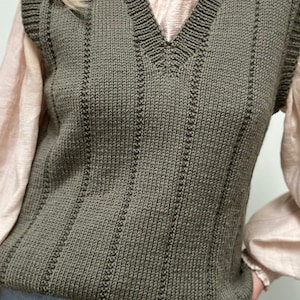 May include: Olive green knitted vest with a V-neck and vertical ribbed detailing. The vest is worn over a light pink blouse and paired with blue jeans. The overall style is casual and layered.