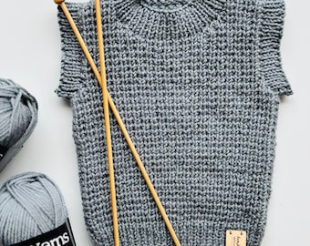 May include: A grey knitted vest with a high collar and short sleeves. Two wooden knitting needles are laid across the vest. Two balls of grey yarn are visible, one with a label that reads "Duch Yarns". A small tag on the vest reads "handmade with love".