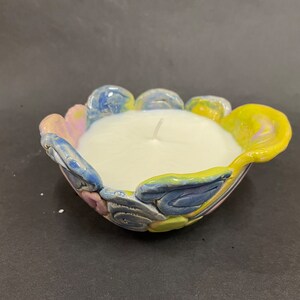 May include: A white candle sits in a colorful ceramic holder. The holder is shaped like a flower with blue, yellow, and pink petals.