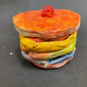 May include: A stack of five ceramic plates in various colors, including red, orange, yellow, green, and blue. The top plate has a small red knob on top.