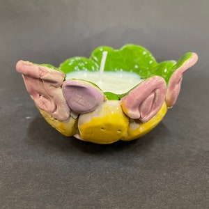 Puede incluir: Un portavelas de cerámica con un diseño floral verde, amarillo, rosa y morado. El portavelas está lleno de una vela blanca.