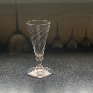 Può includere: Un calice di vetro trasparente con un gambo attorcigliato. Il calice è appoggiato su una superficie grigio scuro.