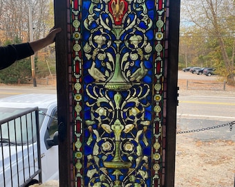 May include: A stained glass window panel with a detailed design of stylized plants, flowers, and birds. The panel is framed in wood and features a variety of colors, including blue, green, red, and gold.