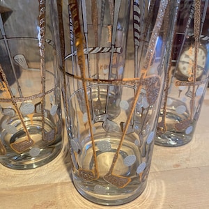 May include: Four clear glass tumblers with gold and white golf club and golf ball designs. The glasses are on a wooden surface.