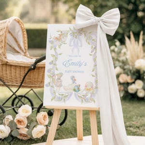 May include: A baby shower sign on a wooden easel, featuring the text "Welcome to Emily's Baby Shower" in elegant script. The sign is decorated with floral and ribbon motifs, and a large white bow adorns the top. A vintage baby carriage and flowers are in the background.