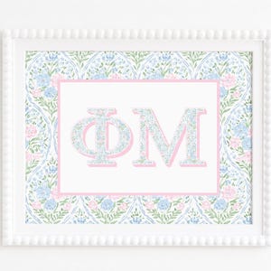Op de afbeelding: Een ingelijste print met de Griekse letters Phi Mu in een bloemenpatroon met blauwe en roze bloemen. De letters zijn gecentreerd op een witte achtergrond met een roze rand, omringd door een herhalend spitsboogpatroon van blauwe en roze bloemen.