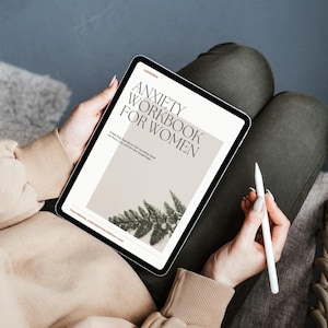 Puede incluir: Una tableta digital que muestra el texto "ANXIETY WORKBOOK FOR WOMEN" con una ilustración de helecho. La tableta está sujeta por una persona que lleva un suéter beige y pantalones verde oliva. También se ve un lápiz óptico blanco.