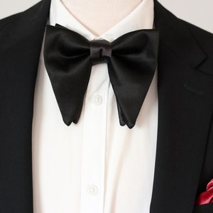May include: A black satin bow tie with a distinctive shape, paired with a white dress shirt and a black suit jacket. A pink pocket square adds a pop of color to the ensemble. Formal wear.