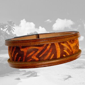 Könnte beinhalten: Ein braunes Lederarmband mit einem orange-braunen, gemusterten Stoffeinsatz. Das Armband ist breit und hat eine abgerundete Form.