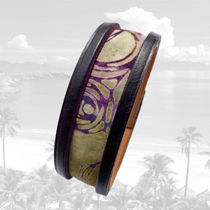 Könnte beinhalten: Ein Armband aus Leder mit violettem und grünem Muster und schwarzem Lederbesatz. Das Armband ist vor einem verschwommenen Hintergrund einer Strandszene zu sehen.