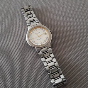 Puede incluir: Un reloj de pulsera plateado y dorado con una esfera blanca y agujas negras. El reloj tiene una correa de metal plateado y las palabras "ELVIA" y "QUARTZ" est&aacute;n impresas en la esfera.