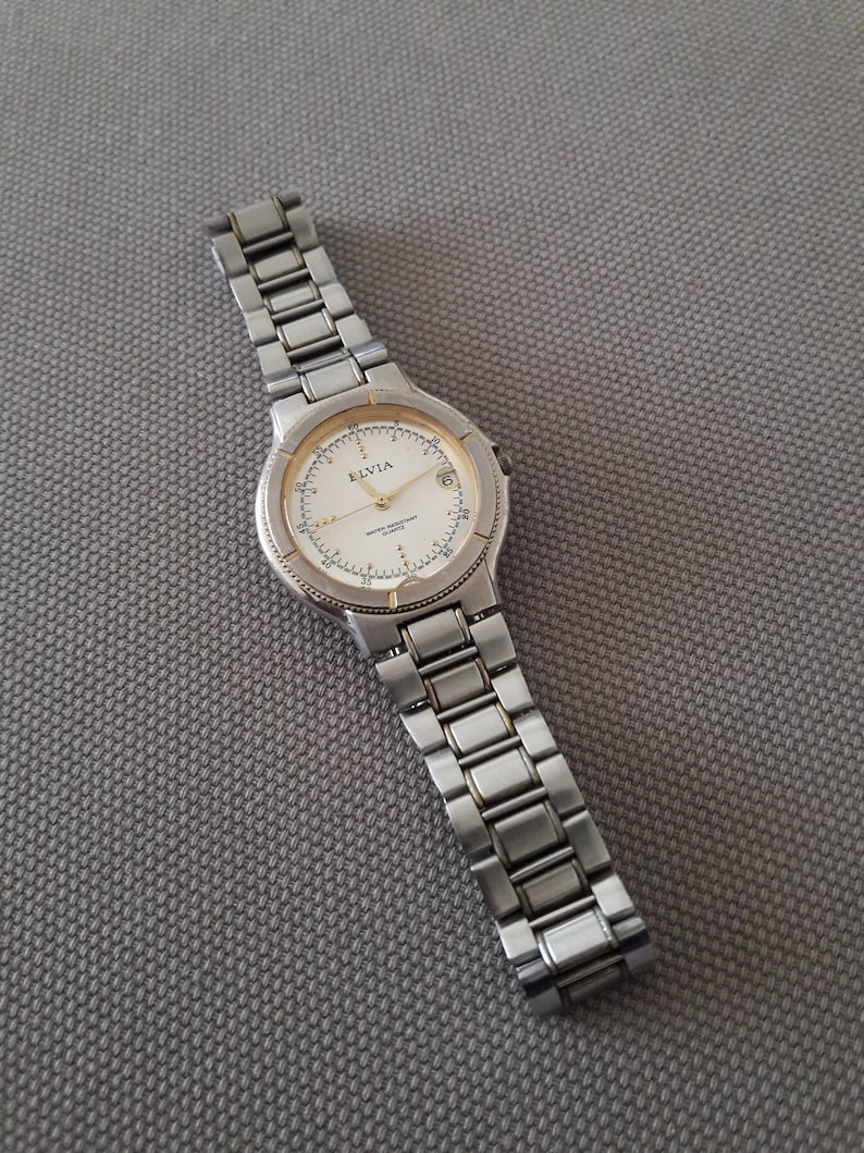 Puede incluir: Un reloj de pulsera plateado y dorado con una esfera blanca y agujas negras. El reloj tiene una correa de metal plateado y las palabras "ELVIA" y "QUARTZ" est&aacute;n impresas en la esfera.