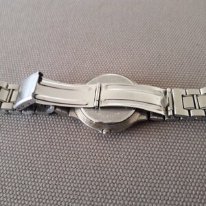 Puede incluir: Un reloj de pulsera plateado con una correa de acero inoxidable. El reloj est&aacute; sobre una superficie gris con la correa doblada hacia atr&aacute;s.